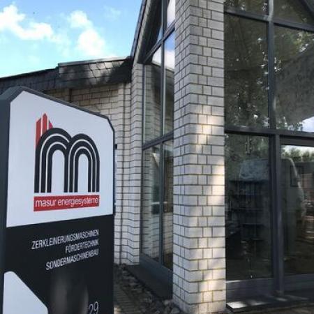 Company sign for Masur Energiesysteme with logo and text referring to shredding machines, conveyor technology, and special machine construction in front of a building with large windows.