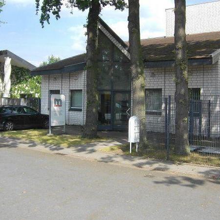 Entrance to a white brick building with a glass front and two trees in front of it, sign with logo and text next to the door
